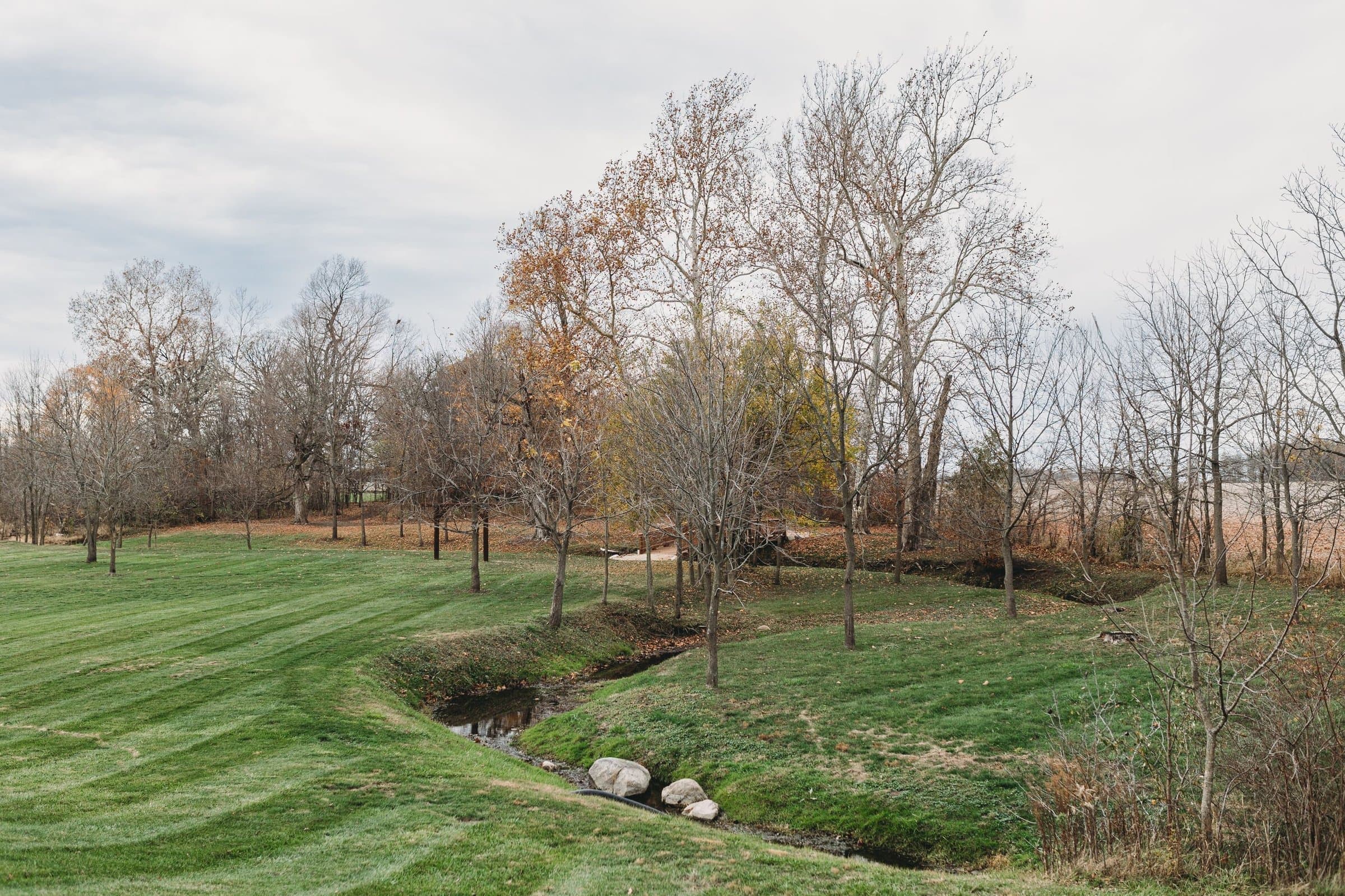 A small creek winding past bare trees and a mowed lawn in late fall